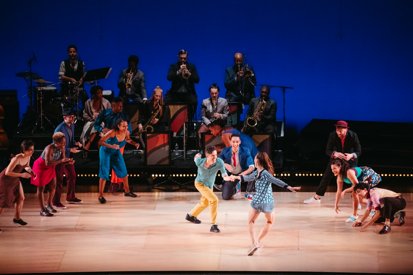 A man and a woman pull away from each other in a classic swing back rock. A semi circle of clapping dancers. A big band plays in the back. 